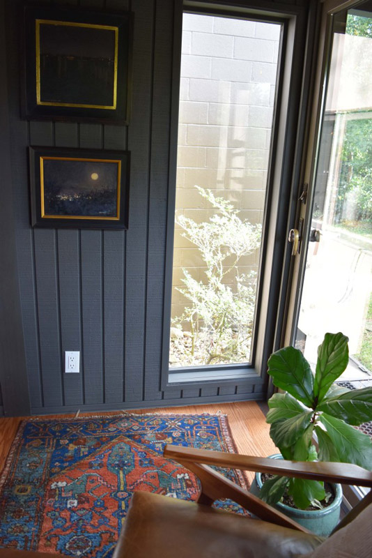 a decorative rug sits next to the glass door entry with framed paintings on the wall nearby, in a reading nook designed by Brand*Eye Home